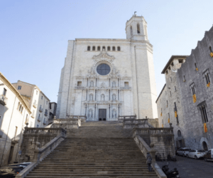 Una amplia escalera de piedra conduce a un imponente edificio histórico blanco con detalles ornamentales y una ventana redonda. Explora el encanto de Girona y su impresionante arquitectura con nuestra aventura "Explora Girona en 48 Horas". - ForTwo - Planes para parejas