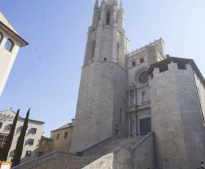 Explora la alta catedral de piedra con intrincados detalles, contrastada por un cielo azul claro, en medio de los encantadores edificios antiguos de Girona en "Explora Girona en 48 Horas". - ForTwo - Planes para parejas