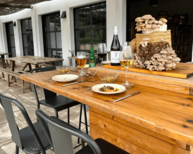Un patio al aire libre cuenta con una mesa de madera para dos, con bebidas, platos de comida y una canasta de las mejores Finca de Setas en Menorca de Menorca como decoración. - ForTwo - Planes para parejas