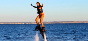 En la vibrante Mallorca, una persona demuestra sus habilidades en el FlyBoard en Mallorca con un traje de baño rojo, volando sobre el océano con una sonrisa radiante y haciendo señales de paz con ambas manos. - ForTwo - Planes para parejas