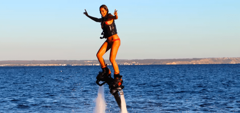 En la vibrante Mallorca, una persona demuestra sus habilidades en el FlyBoard en Mallorca con un traje de baño rojo, volando sobre el océano con una sonrisa radiante y haciendo señales de paz con ambas manos. - ForTwo - Planes para parejas