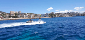 Alguien está disfrutando del FlyBoard en Mallorca sobre un océano resplandeciente, con una moto acuática cerca y edificios de la ciudad elevándose en la distancia. - ForTwo - Planes para parejas