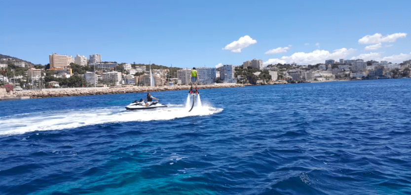 Alguien está disfrutando del FlyBoard en Mallorca sobre un océano resplandeciente, con una moto acuática cerca y edificios de la ciudad elevándose en la distancia. - ForTwo - Planes para parejas