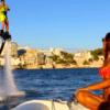Utilizando un FlyBoard en Mallorca, una persona se eleva sobre el mar centelleante, cautivando la atención bajo el cielo azul claro mientras una mujer en bikini observa desde un barco cercano. - ForTwo - Planes para parejas