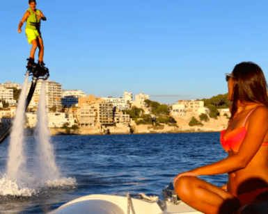 Utilizando un FlyBoard en Mallorca, una persona se eleva sobre el mar centelleante, cautivando la atención bajo el cielo azul claro mientras una mujer en bikini observa desde un barco cercano. - ForTwo - Planes para parejas