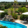 Relájese junto a la piscina del Hotel Rural en Menorca, con tumbonas, sombrillas, exuberante vegetación y árboles lejanos bajo el cielo despejado. - ForTwo - Planes para parejas