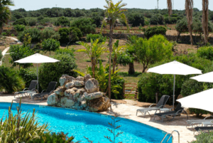 Relájese junto a la piscina del Hotel Rural en Menorca, con tumbonas, sombrillas, exuberante vegetación y árboles lejanos bajo el cielo despejado. - ForTwo - Planes para parejas