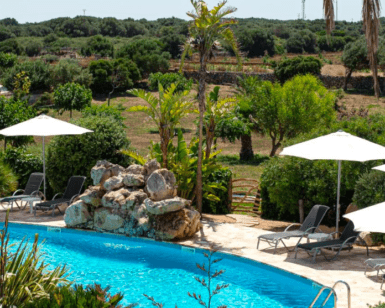Relájese junto a la piscina del Hotel Rural en Menorca, con tumbonas, sombrillas, exuberante vegetación y árboles lejanos bajo el cielo despejado. - ForTwo - Planes para parejas