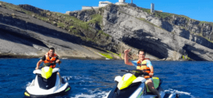 Dos personas en "Jet Ski por la Costa de Cantabria" exploran las vibrantes aguas cerca de los acantilados rocosos mientras usan chalecos salvavidas. - ForTwo - Planes para parejas