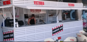 La barra exterior blanca y roja, con el letrero Paddock Bar, captura el ambiente animado de Málaga. Una persona trabaja activamente en el interior, lo que lo convierte en el lugar perfecto para relajarse después de un emocionante día en Kart Extreme en Málaga. - ForTwo - Planes para parejas