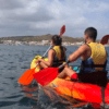 Dos kayakistas con chalecos salvavidas navegan por el mar cerca de Menorca, con un pueblo costero y colinas detrás, mostrando la tranquila belleza de "Kayak por la Reserva Natural de Menorca. - ForTwo - Planes para parejas