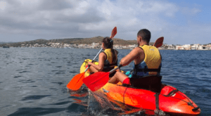 Dos kayakistas con chalecos salvavidas navegan por el mar cerca de Menorca, con un pueblo costero y colinas detrás, mostrando la tranquila belleza de "Kayak por la Reserva Natural de Menorca. - ForTwo - Planes para parejas