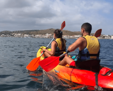 Dos kayakistas con chalecos salvavidas navegan por el mar cerca de Menorca, con un pueblo costero y colinas detrás, mostrando la tranquila belleza de "Kayak por la Reserva Natural de Menorca. - ForTwo - Planes para parejas