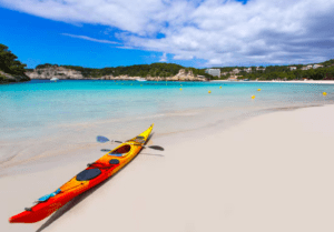 A Kayak por la Reserva Natural de Menorca se encuentra en una playa de arena, entre aguas turquesas cristalinas y un cielo parcialmente nublado, mostrando la impresionante belleza de la zona. - ForTwo - Planes para parejas