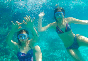 En las cristalinas aguas de Menorca, dos personas con snorkel y traje de baño nadan bajo el agua cerca de una pintoresca Reserva Natural, con la luz del sol filtrándose a través de ellas. - ForTwo - Planes para parejas