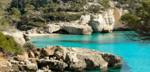 Aguas turquesas y acantilados rocosos rodean una exuberante vegetación en una serena escena costera, haciéndose eco de la belleza intacta que se experimenta con la "Lancha Sin Licencia en Menorca". - ForTwo - Planes para parejas