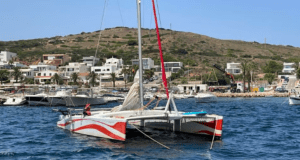 La "Navega en Catamarán por Menorca", con detalles en rojo, está anclada en un puerto deportivo de Menorca con un telón de fondo de colinas y edificios. - ForTwo - Planes para parejas