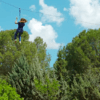 Deslizarse en tirolina entre frondosos árboles verdes contra un cielo azul brillante en el Parque de Aventura en Hornachos encapsula la emoción de la aventura. - ForTwo - Planes para parejas