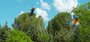 Deslizarse en tirolina entre frondosos árboles verdes contra un cielo azul brillante en el Parque de Aventura en Hornachos encapsula la emoción de la aventura. - ForTwo - Planes para parejas