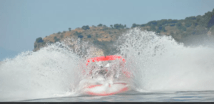 Paseo de Acrobacias en el Agua en Girona" presenta una lancha rápida que crea grandes salpicaduras a medida que avanza rápidamente por el agua, con un telón de fondo de colinas pintorescas que evocan Girona. - ForTwo - Planes para parejas