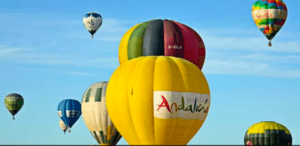 Una vibrante escena de coloridos globos aerostáticos, incluido uno grande amarillo con la palabra "Andalucía", decora el cielo azul claro, evocando la encantadora experiencia de un "Paseo en Globo de Almodóvar del Río". - ForTwo - Planes para parejas