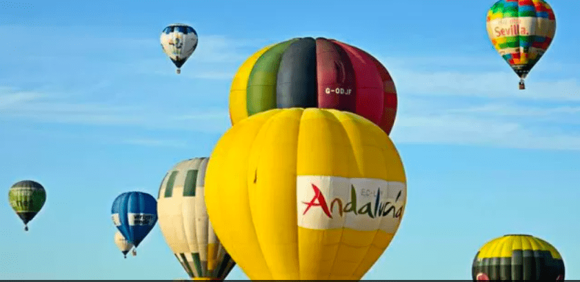 Una vibrante escena de coloridos globos aerostáticos, incluido uno grande amarillo con la palabra "Andalucía", decora el cielo azul claro, evocando la encantadora experiencia de un "Paseo en Globo de Almodóvar del Río". - ForTwo - Planes para parejas