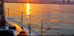 El "Paseo exclusivo en Velero en la Costa Brava" ofrece una impresionante vista de la puesta de sol sobre un mar en calma, enmarcada por las siluetas del horizonte de la ciudad, ideal para sumergirse en la belleza del horizonte de la Costa Brava. - ForTwo - Planes para parejas