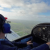 En la cabina de un pequeño avión, una persona disfruta de la emoción de ser Piloto por un Día en Córdoba, con unos auriculares y una gorra azules mientras vuela sobre el paisaje rural en un día claro. - ForTwo - Planes para parejas