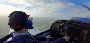 En la cabina de un pequeño avión, una persona disfruta de la emoción de ser Piloto por un Día en Córdoba, con unos auriculares y una gorra azules mientras vuela sobre el paisaje rural en un día claro. - ForTwo - Planes para parejas