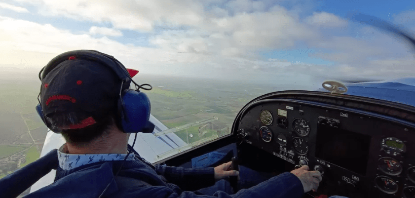 En la cabina de un pequeño avión, una persona disfruta de la emoción de ser Piloto por un Día en Córdoba, con unos auriculares y una gorra azules mientras vuela sobre el paisaje rural en un día claro. - ForTwo - Planes para parejas