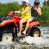 Dos personas experimentan un emocionante Quad por el río Guadalhorce, conduciendo un todoterreno rojo por las aguas poco profundas del río, entre una exuberante vegetación verde. - ForTwo - Planes para parejas