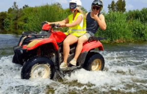 Dos personas experimentan un emocionante Quad por el río Guadalhorce, conduciendo un todoterreno rojo por las aguas poco profundas del río, entre una exuberante vegetación verde. - ForTwo - Planes para parejas