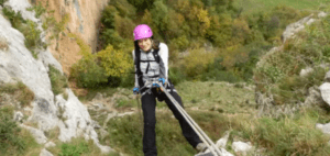 Con un casco rosa, una persona sube hábilmente una pendiente rocosa con cuerdas, acercándose a la entrada de Cueva Mur para una aventura de Rápel en Cueva Mur. - ForTwo - Planes para parejas