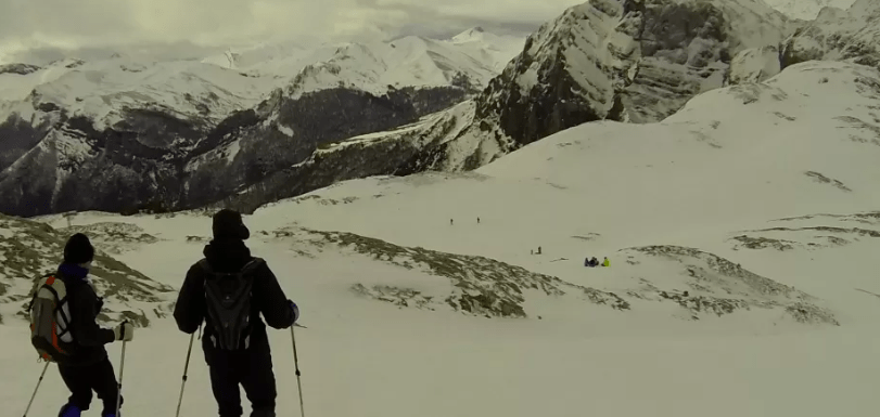 Dos personas esquían por una montaña nevada con nubes y picos lejanos, que recuerdan a los majestuosos Picos de Europa, similar a la experiencia que ofrecen Raquetas de Nieve en Picos de Europa. - ForTwo - Planes para parejas