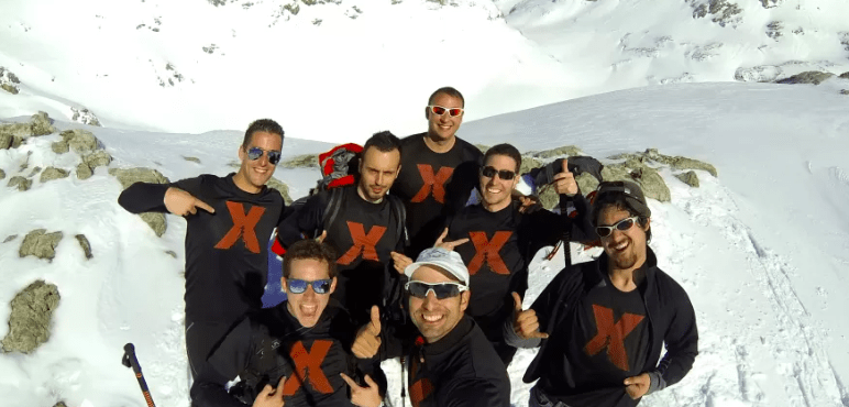 Siete personas con atuendos iguales sostienen bastones de esquí de Raquetas de Nieve en Picos de Europa y llevan gafas de sol, posando en la ladera de una montaña nevada y sonriendo. - ForTwo - Planes para parejas