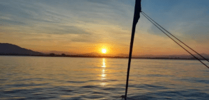 Una puesta de sol sobre aguas tranquilas, montañas recortadas detrás y el elegante casco de un catamarán al frente preparan el escenario para un recorrido romántico en catamarán. - ForTwo - Planes para parejas