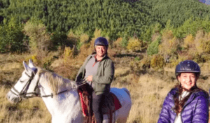 Dos individuos con casco, uno sobre un caballo blanco y otro de pie, disfrutan de la pintoresca Ruta a Caballo en Villanovilla en medio de las verdes colinas. - ForTwo - Planes para parejas
