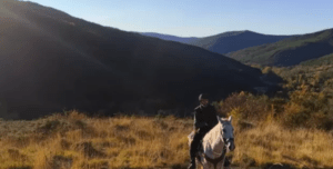 Una persona emprende la Ruta a Caballo en Villanovilla, montado en un caballo blanco a través de colinas cubiertas de hierba con las majestuosas montañas de Villanovilla elevándose bajo un cielo despejado. - ForTwo - Planes para parejas