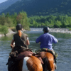 Dos personas disfrutan de una "Ruta a Caballo en Villanovilla", cabalgando a lo largo de un pintoresco río con un exuberante bosque y majestuosas montañas como telón de fondo. - ForTwo - Planes para parejas