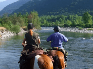 Dos personas disfrutan de una "Ruta a Caballo en Villanovilla", cabalgando a lo largo de un pintoresco río con un exuberante bosque y majestuosas montañas como telón de fondo. - ForTwo - Planes para parejas