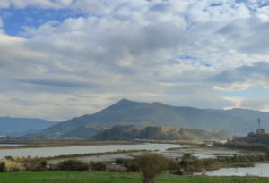 El cielo nublado cubre las majestuosas montañas, un río serpentea entre los campos en primer plano, evocando la esencia de "Sabor y tradición en los Valles Pasiegos", donde la belleza de la naturaleza se encuentra con la riqueza cultural. - ForTwo - Planes para parejas
