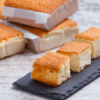 Cuatro trozos de bizcocho brillan con un encanto dorado sobre una pizarra, evocando la deliciosa tradición del "Sabor y tradición en los Valles Pasiegos", mientras pasteles enteros envueltos en papel se yerguen majestuosos en el fondo, celebrando la herencia culinaria. - ForTwo - Planes para parejas