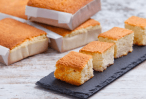 Cuatro trozos de bizcocho brillan con un encanto dorado sobre una pizarra, evocando la deliciosa tradición del "Sabor y tradición en los Valles Pasiegos", mientras pasteles enteros envueltos en papel se yerguen majestuosos en el fondo, celebrando la herencia culinaria. - ForTwo - Planes para parejas