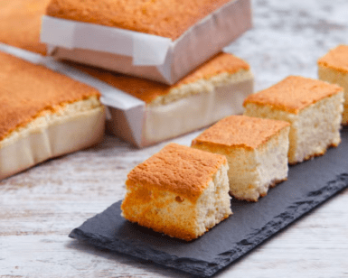 Cuatro trozos de bizcocho brillan con un encanto dorado sobre una pizarra, evocando la deliciosa tradición del "Sabor y tradición en los Valles Pasiegos", mientras pasteles enteros envueltos en papel se yerguen majestuosos en el fondo, celebrando la herencia culinaria. - ForTwo - Planes para parejas