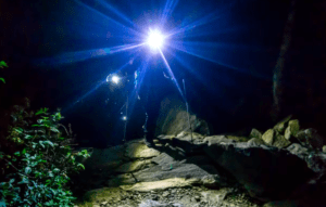 Con una linterna frontal brillante y bastones de trekking, el excursionista navega por el paisaje rocoso rodeado de exuberante vegetación, que recuerda a las misteriosas cuevas de Menorca durante una aventura de Senderismo Nocturno en Menorca. - ForTwo - Planes para parejas