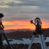 Dos trípodes sostienen telescopios apuntando a una vibrante puesta de sol menorquina sobre colinas distantes, cerca de un muro de piedra: un preludio perfecto para una noche de Senderismo Nocturno en Menorca bajo las estrellas. - ForTwo - Planes para parejas