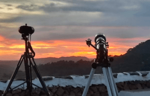 Dos trípodes sostienen telescopios apuntando a una vibrante puesta de sol menorquina sobre colinas distantes, cerca de un muro de piedra: un preludio perfecto para una noche de Senderismo Nocturno en Menorca bajo las estrellas. - ForTwo - Planes para parejas