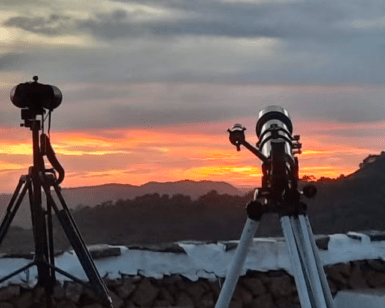 Dos trípodes sostienen telescopios apuntando a una vibrante puesta de sol menorquina sobre colinas distantes, cerca de un muro de piedra: un preludio perfecto para una noche de Senderismo Nocturno en Menorca bajo las estrellas. - ForTwo - Planes para parejas