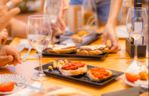 Manos que se acercan a los aperitivos en una mesa con copas de vino, aceitunas y delicias de Visita a la Quesería Subaida. - ForTwo - Planes para parejas
