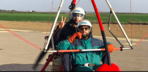 Dos personas en una Vuela el Guadalquivir con Ala Delta Motorizada, ataviadas con cascos y gafas protectoras, se preparan para despegar desde un aeródromo con exuberante hierba, listas para sobrevolar paisajes escénicos que recuerdan al Guadalquivir. - ForTwo - Planes para parejas